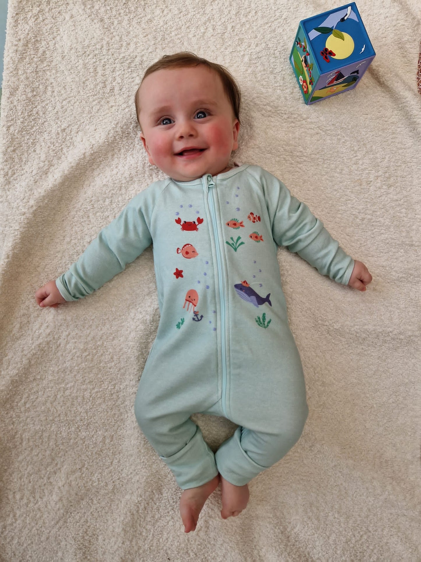 Bébé allongé sur tapis et souriant portant le pyjama "nager au lieu des coraux" de la collection mer.